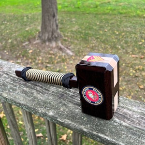 Può includere: Un mazzuolo di legno con un manico a strisce nere e beige. Il mazzuolo ha una finitura in legno scuro e un emblema del Corpo dei Marines degli Stati Uniti sul davanti. L'emblema è rosso, bianco e blu con un'aquila dorata e le parole "United States Marine Corps".