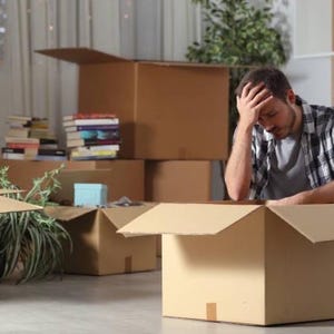 May include: A person sits on the floor surrounded by cardboard boxes, looking overwhelmed and stressed. The boxes are packed with belongings, suggesting a move or relocation.