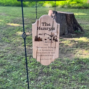Könnte beinhalten: Ein Holzschild mit dem Text "The Henry's" und "The Drunkest Bunch of Assholes This Side of the Campground!" hängt an einem schwarzen Metall-Gartenstecker. Das Schild hat ein Camping-Thema mit einer Berg- und Wohnwagen-Grafik. Das Schild befindet sich in einer grasbewachsenen Umgebung im Freien.