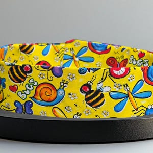 May include: A yellow fabric storage bin with a colourful bug print. The bin features a variety of bugs, including butterflies, ladybugs, bees, snails, and dragonflies. The bin has the word "bugs" printed on it.