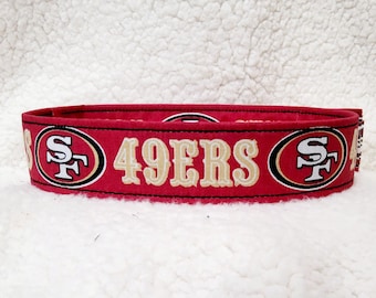 以下が含まれることがあります： 金色の文字で「49ERS」と書かれた赤い生地の首輪。首輪の両側にチームのロゴがあり、黒い縁取りがされています。背景は白のテクスチャード加工された生地です。