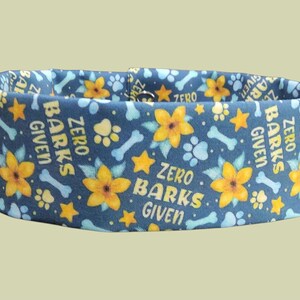 May include: A blue dog collar with a repeating pattern of yellow flowers, stars, paw prints, and bones. The text "ZERO BARKS GIVEN" is printed on the collar. The collar is designed for pets.