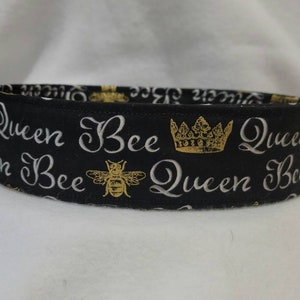 May include: Black fabric headband with a white and gold print of the words "Queen Bee" and a crown. The headband is decorated with a gold bee.