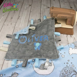 May include: A grey and blue baby blanket with a personalised name "Oliver" and an embroidered elephant. The blanket has multiple satin ribbons attached to the edges.