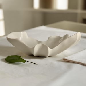 Könnte beinhalten: Eine cremefarbene Keramikschale mit organischer Form und welligem Rand. Die Schale steht auf einer weißen Oberfläche, daneben ein grünes Blatt und eine gefaltete weiße Serviette. Das Design der Schale ist modern und minimalistisch.