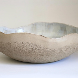 Może przedstawiać: Ręcznie wykonana, beżowa ceramiczna miska o teksturowanej powierzchni zewnętrznej i gładkim, jasnym wnętrzu. Miska ma organiczny, falisty brzeg i zaokrąglony kształt, idealna do serwowania lub ekspozycji.