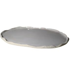 Puede incluir: Una bandeja de servicio ovalada de cerámica con una superficie lisa y brillante esmaltada en gris claro. Los bordes de la bandeja son irregulares y texturizados, con un borde de arcilla mate, color blanco roto, sin acabado.