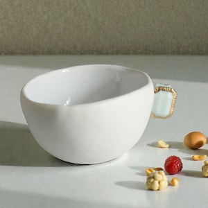 Puede incluir: Una taza de té de cerámica blanca con un diseño asimétrico único. El asa es de color azul claro con detalles dorados. Alrededor de la taza hay pequeños bocadillos, como cacahuetes, bayas y palomitas de maíz.
