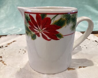 Könnte beinhalten: Ein weißer Keramik-Milchkännchen mit einem roten Weihnachtsstern- und grünem Blattwerk-Design. Der Milchkännchen hat einen Griff und einen Ausguss.