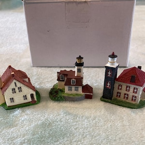 Può includere: Tre modelli di fari in miniatura, ognuno con una torre bianca e un tetto rosso, sono esposti su una superficie verde. I fari sono in ceramica e presentano dettagli intricati, tra cui finestre e porte.