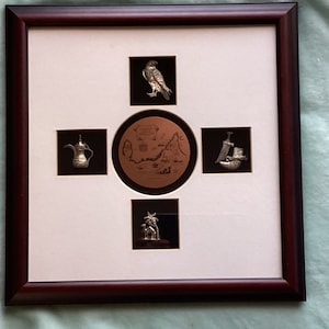 May include: A framed artwork with five silver-toned metal plaques depicting various cultural symbols. The center plaque features a map of a desert landscape with the text "United Arab Emirates". The other plaques depict a falcon, a coffee pot, a dagger, and a camel with a palm tree.