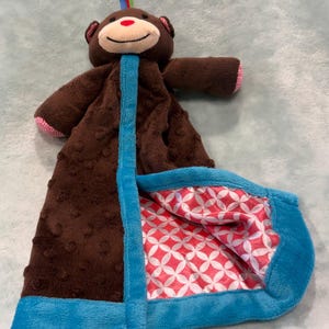Puede incluir: Una manta de seguridad para bebé con forma de mono de peluche marrón, con una cara sonriente, ribetes azules y un interior estampado. La manta presenta un exterior suave y texturizado y un diseño colorido en el interior.