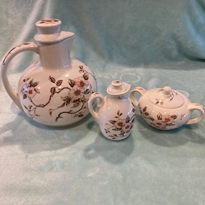 May include: A set of three white ceramic tea or coffee serving pieces with a floral design. The pieces include a pitcher, a creamer, and a sugar bowl.
