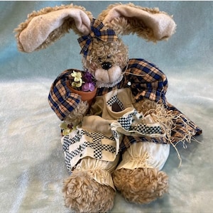 May include: A plush brown bunny wearing a blue and yellow plaid shirt and a white apron with black and white checkered trim. The bunny is holding a small pot of purple flowers.