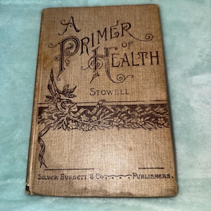 Puede incluir: Un libro antiguo titulado "A Primer of Health" de Stowell. La portada es de color marrón claro con letras ornamentadas en marrón oscuro e ilustraciones florales. El nombre del editor, Silver, Burdett & Co., está impreso en la parte inferior.