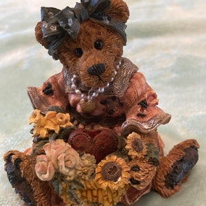 May include: A brown teddy bear figurine wearing a pink dress with a blue bow and a pearl necklace. The bear is sitting on a bed of sunflowers and other flowers.