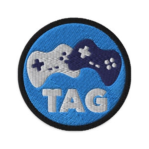 Pode incluir: Um remendo bordado azul e preto com um controlador de videogame branco e roxo e a palavra "TAG" em branco.