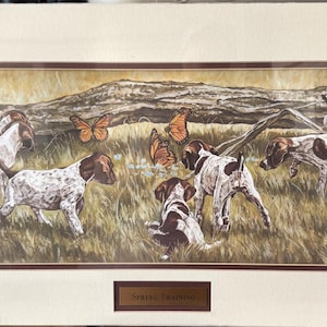 May include: A framed print depicting a painting of dogs with brown and white markings in a grassy field. The dogs are interacting with orange butterflies near a fallen log. The artwork is framed with a brown border and a cream-coloured mount.