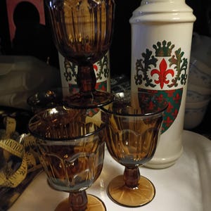 May include: A collection of amber-colored glassware, including stemmed goblets and smaller wine glasses. A white ceramic bottle with a decorative emblem featuring a fleur-de-lis is also present. The glassware is arranged on a white surface.