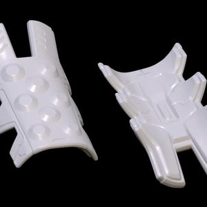 May include: Two white plastic pieces of armor for a costume. One piece has a textured surface with raised circles. The other piece has a more angular design.