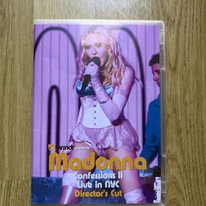 Op de afbeelding: Een dvd-hoes met een concertoptreden van Madonna. De hoes toont een blonde artiest in een wit korset en kanten rok die een microfoon vasthoudt. De tekst op de hoes luidt: Grindr presents Madonna Confessions II Live in NYC Director's Cut.