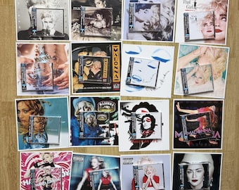 May include: A collection of twelve Madonna music albums, each featuring a square vinyl record sleeve paired with its corresponding compact disc. The display showcases various iconic portraits and graphic designs from the artist's discography.
