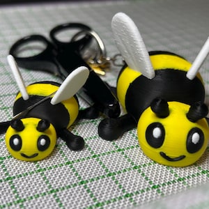 May include: Two 3D-printed bee keyrings. The bees are yellow with black stripes, black legs, and white wings. Each bee has a smiling face with black eyes and a black mouth. A pair of black scissors and a keyring are visible in the background.