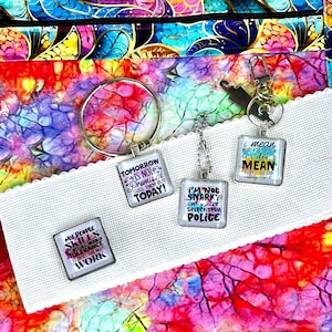 May include: A collection of keychains with humorous quotes on a colorful patterned background. The keychains have square pendants with phrases like "Tomorrow is not promised, cuss them out today!" and "I'm not snarky, I'm just selectively polite."
