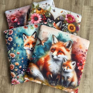May include: A collection of decorative throw pillows featuring watercolor illustrations of foxes and floral arrangements. The pillows showcase vibrant colors, including orange, red, and blue, with detailed depictions of foxes and various flowers.