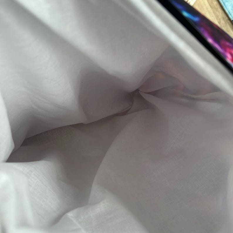 May include: Interior view of a white fabric bag with a colorful galaxy print on the outer edge. The bag's interior is a soft, white material, creating a contrast with the vibrant outer design. The bag appears to be empty.