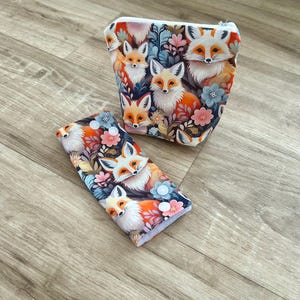 May include: Two-piece set featuring a cosmetic bag and a matching case, both adorned with a whimsical fox and floral pattern. The design showcases foxes in shades of orange, white, and brown, set against a backdrop of flowers and foliage. The cosmetic bag has a zipper closure.