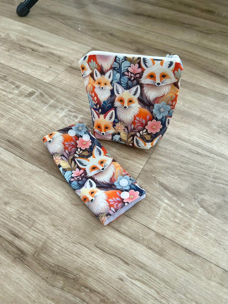 May include: Two-piece set featuring a cosmetic bag and a matching case, both adorned with a whimsical fox and floral pattern. The design showcases foxes in shades of orange, white, and brown, set against a backdrop of flowers and foliage. The cosmetic bag has a zipper closure.