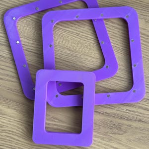 May include: Three square, purple frames with rounded corners and small, silver-coloured circular accents. The frames are of varying sizes, with the smallest one in the foreground. The frames are arranged on a light brown wooden surface.