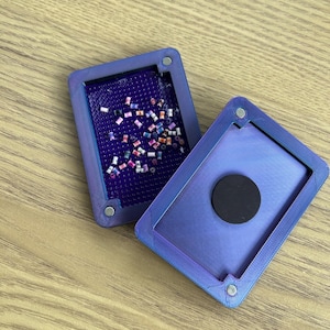 Peut inclure: Un organisateur de perles rectangulaire, violet iridescent, avec un couvercle à charnière. L'organisateur ouvert révèle un plateau rempli de petites perles colorées. Le couvercle a un aimant circulaire noir. L'organisateur est sur une surface en bois.