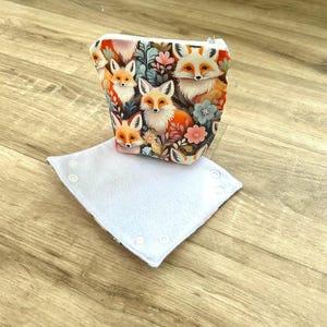 May include: A small, zippered pouch with a vibrant fox and floral print. The pouch is white with a colorful design of foxes, flowers, and foliage. A white, square, buttoned item is in front of the pouch.