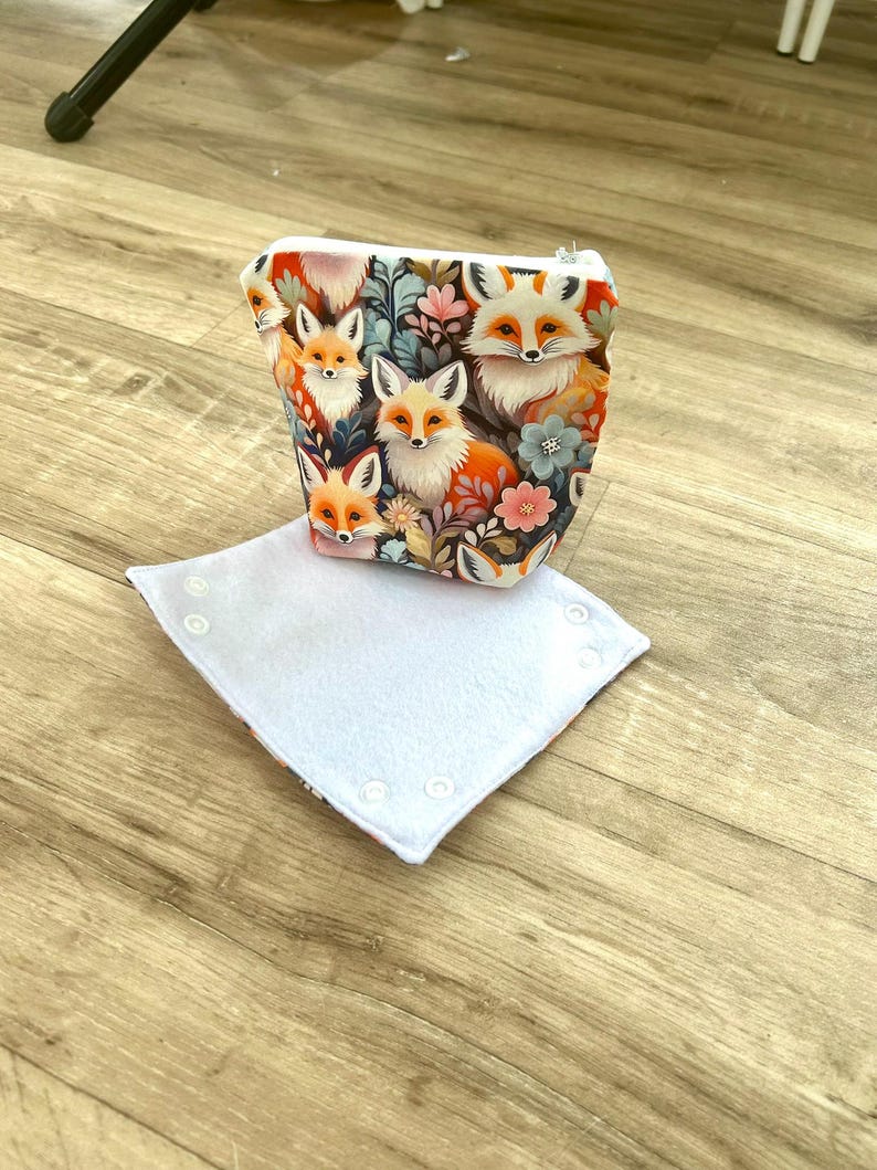 May include: A small, zippered pouch with a vibrant fox and floral print. The pouch is white with a colorful design of foxes, flowers, and foliage. A white, square, buttoned item is in front of the pouch.