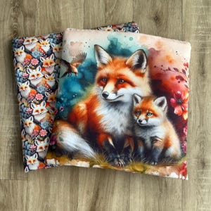 May include: Two decorative throw pillows. One pillow features a repeating pattern of foxes and flowers. The other pillow displays a watercolor painting of a mother fox and her cub, with a bird in flight and floral accents.