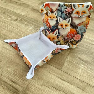 May include: A small, open tray and a zippered pouch featuring a repeating pattern of foxes and floral elements. The foxes have orange and white fur, set against a backdrop of dark blue, pink, and green. The tray has a white interior.