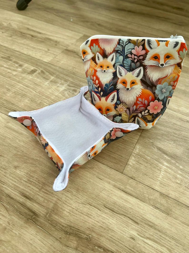 May include: A small, open tray and a zippered pouch featuring a repeating pattern of foxes and floral elements. The foxes have orange and white fur, set against a backdrop of dark blue, pink, and green. The tray has a white interior.