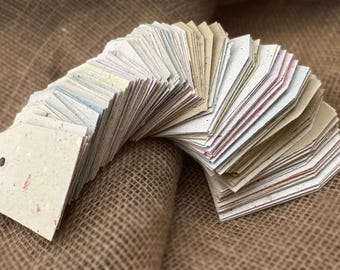 May include: A stack of handmade paper tags in various shades of beige, cream, and light brown. The tags are arranged in a semi-circle and have a rustic, handmade appearance.