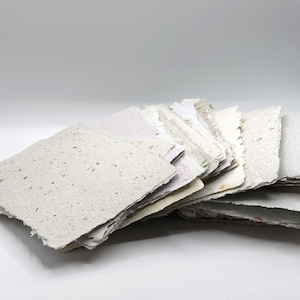 May include: A stack of rectangular handmade paper sheets with rough, deckled edges. The paper is off-white and light gray, featuring visible organic fiber inclusions and flecks throughout the texture of each individual sheet in the collection.