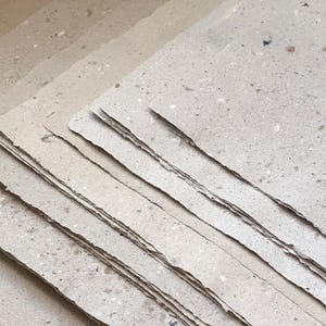 May include: A stack of handmade paper sheets with deckled edges. The paper has a speckled texture with a light beige color. The sheets are layered, showing the varying tones of the paper. Ideal for art projects or stationery.