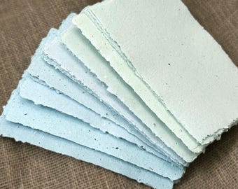 May include: A stack of handmade paper sheets in shades of blue and green. The paper has a textured surface and irregular, torn edges. The sheets are stacked on a brown, textured surface.