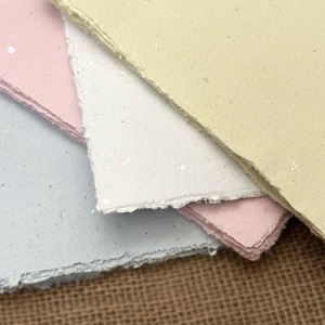 May include: A stack of handmade paper sheets in various pastel colors: pale yellow, pink, white, and light blue. The paper has a textured surface and deckled edges, suggesting a craft or art supply.