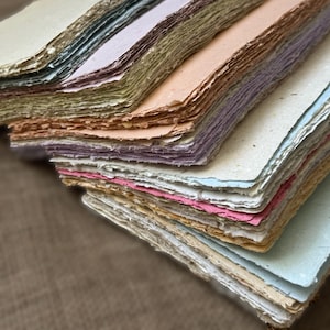 May include: A stack of rectangular handmade paper sheets with rough, deckled edges. The paper comes in a variety of soft pastel colors including cream, light blue, pale pink, lavender, peach, and mint green, featuring a visible fibrous texture.