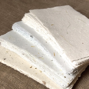 May include: A stack of rectangular, off-white handmade paper sheets with deckled edges. The paper has a textured, fibrous surface containing visible small organic inclusions and plant fibers embedded throughout the light cream-colored material.