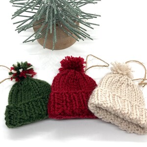 May include: Three knitted Christmas ornaments shaped like winter hats. The hats are green, red, and beige. Each hat has a white pom-pom on top.