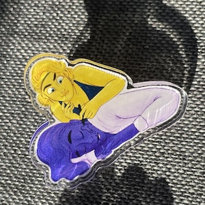 May include: A clear acrylic charm featuring a cartoon illustration of two people. One person has yellow hair and is wearing a blue and yellow outfit. The other person has purple hair and is wearing a purple and white outfit. The person with yellow hair is holding the other person's hand.