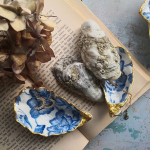 Könnte beinhalten: Zwei blaue und weiße Porzellan-Austernmuscheln mit goldenen Verzierungen, die als dekorative Schalen verwendet werden. Die Muscheln sind mit Schmuck gefüllt, darunter eine goldene Halskette und ein Paar Ohrringe. Die Muscheln liegen auf einem aufgeschlagenen Buch mit Text in deutscher Sprache.