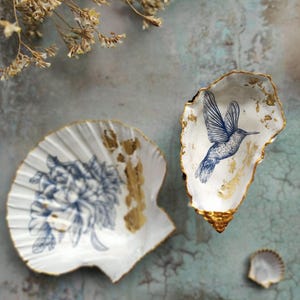 Könnte beinhalten: Zwei weiße Muschelschalen mit goldenen Akzenten. Eine Schale zeigt ein blaues Blumendesign, die andere einen blauen Kolibri. Eine kleine Muschel ist ebenfalls vorhanden. Die Schalen befinden sich auf einer türkis-grauen Oberfläche.